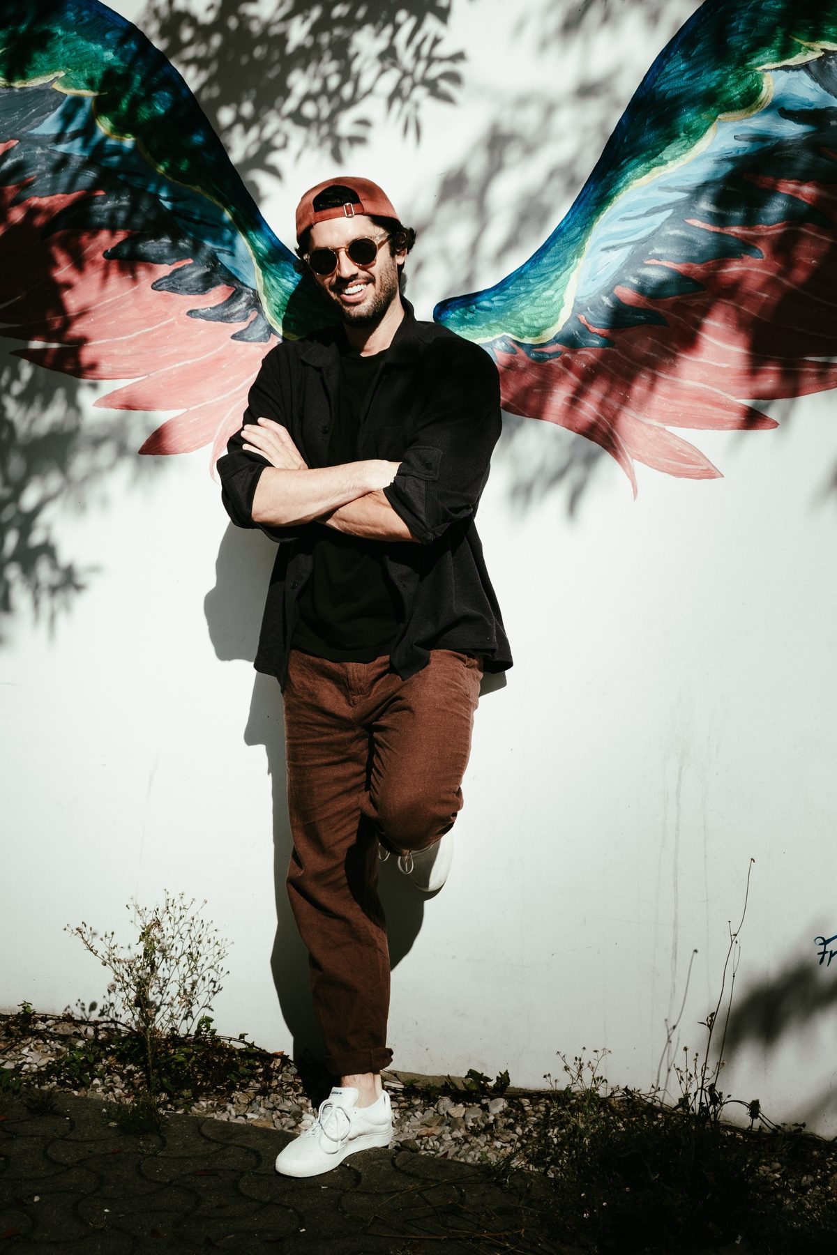 Editorial-Portrait vor einem bunten Flügel-Wandbild, lachender Mann mit Sonnenbrille und Cap